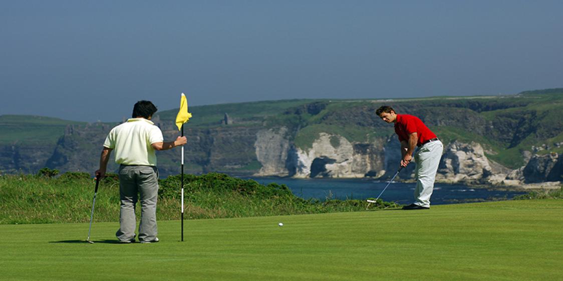 Two men putting on the golf green in Royal Portrush, with background of ocean views and mountains.