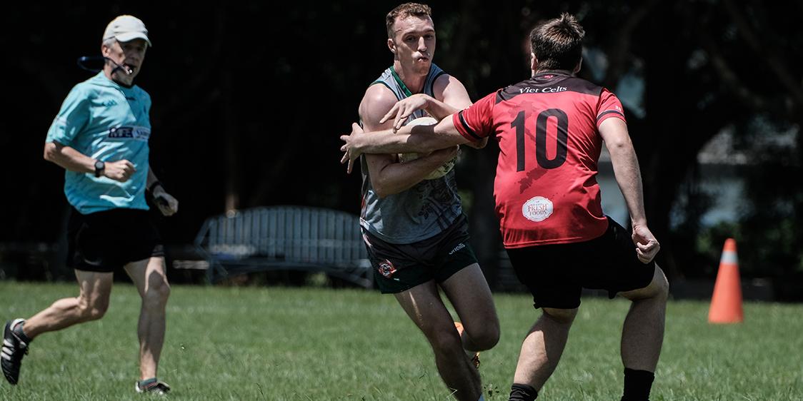 A Hong Kong player in a grey and green jersey holds the ball while attempting to evade or tackle a Viet Celts player wearing a red and black jersey with the number 10. The action takes place on a grassy field with trees in the background. A person in a light blue shirt and black shorts, possibly a referee, is visible behind the players. The image captures a dynamic moment of athletic competition between the two teams.