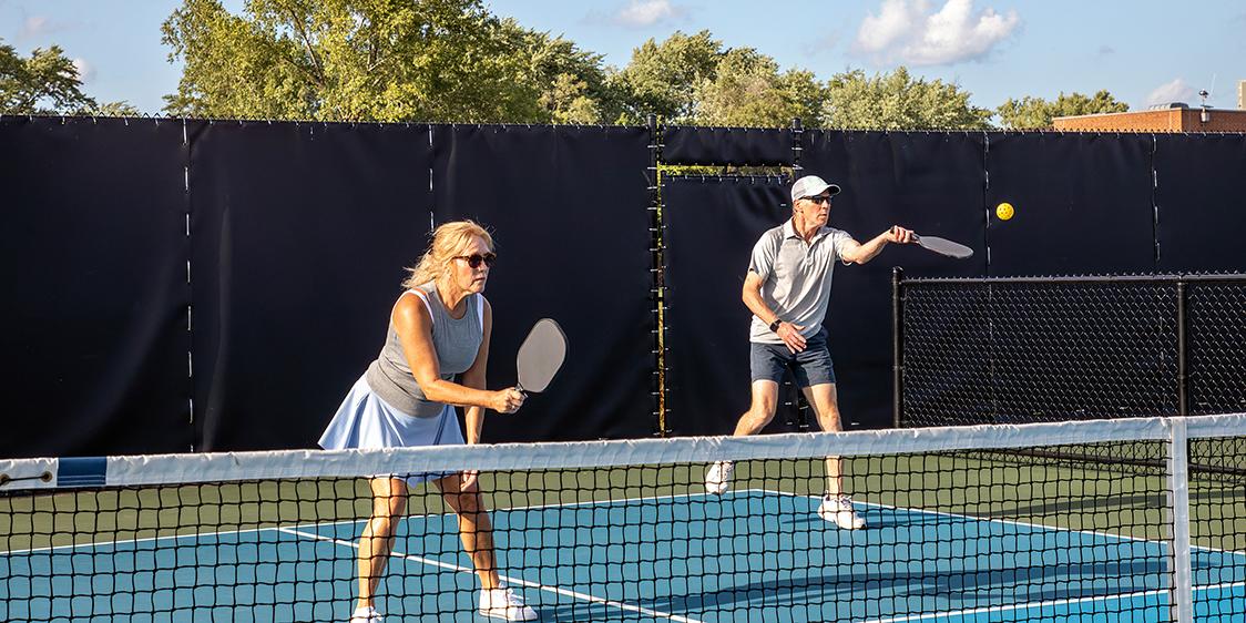 Two people playing pickleball on an outdoor court with trees and a building in the background
