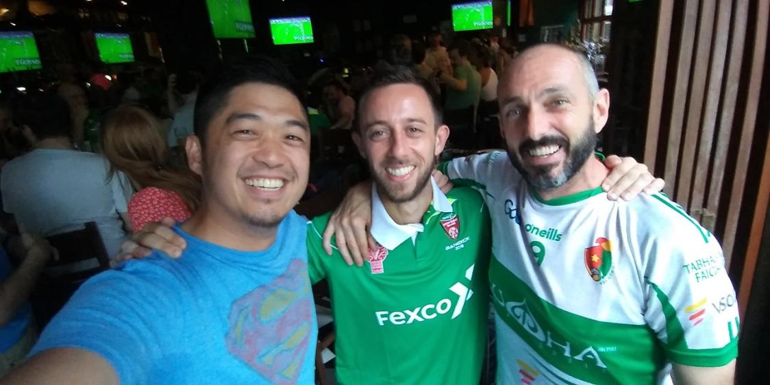 Three people standing together in a bar, smiling at the camera. The person on the left wears a blue Superman shirt, while the two on the right wear green sports jerseys with logos including 'Fexco' and 'O'Neills'. Multiple TV screens in the background display a sports game, suggesting a celebratory or social sporting event atmosphere
