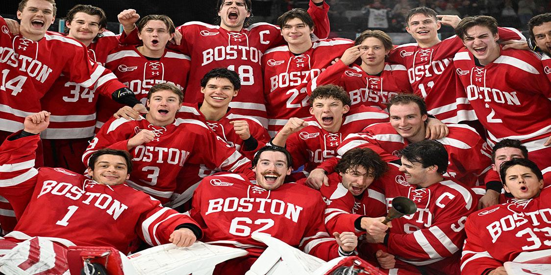 Group photo of winners of the Friendship Four 2024, Boston University ice hockey team wearing matching red uniforms with white stripes and the word “BOSTON” printed across the chest. 