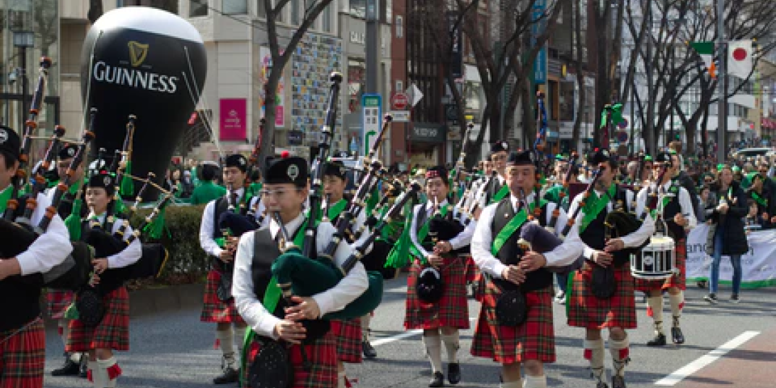 Marching band playing bagpipes during a St Patrick’s Day parade in Tokyo, wearing green clothing and tartan kilts as they move through a busy urban street.