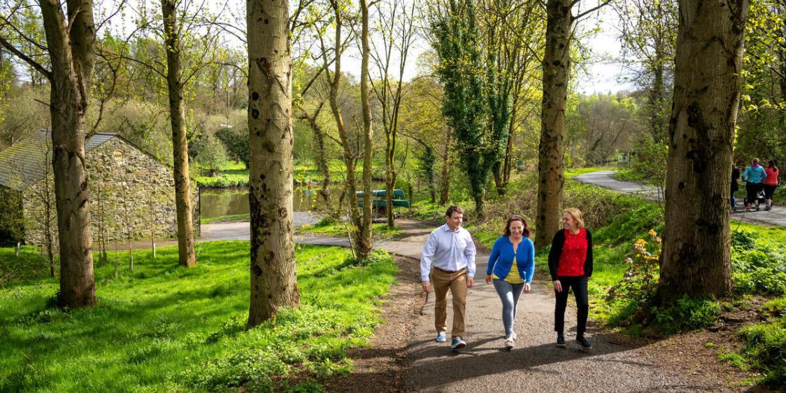 Three people walking along a tree‑lined riverside path on a sunny day, with spring greenery and other walkers in the background