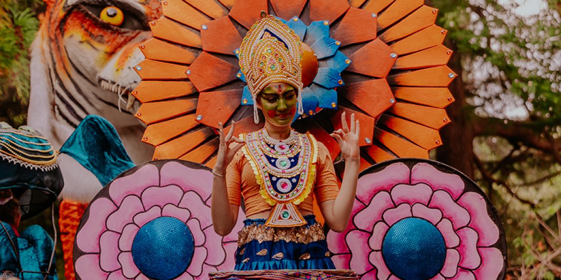 Performer in a vibrant costume at a cultural festival, with a giant tiger figure behind.  Provide your feedback on BizChat