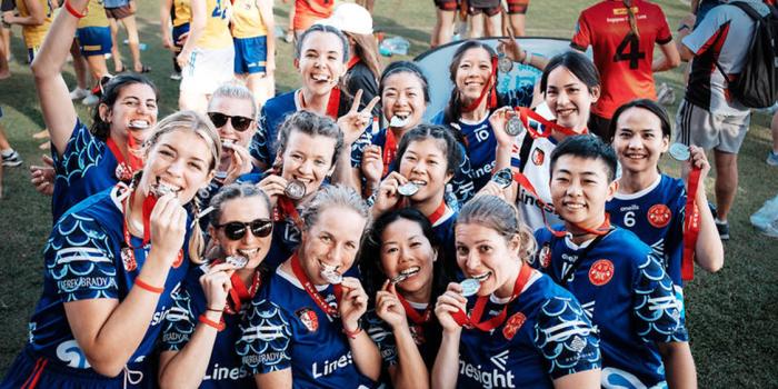 Members of The Taiwan Celts Gaelic Football Club’s women’s team wearing blue sports jerseys with white patterns and red medals around their necks, gathered closely together on a grassy field. They are smiling, holding up their medals, with some making peace signs and others playfully biting their medals. Other athletes in sports attire are visible in the background